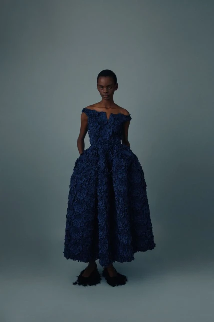 Elegant woman wearing a dark blue, off-the-shoulder textured gown standing indoors against a gray backdrop in a studio shot.