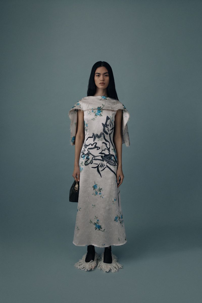 Elegant woman poses in a patterned dress with oversized collar, carrying a black purse, with unique fuzzy footwear against a plain blue backdrop.