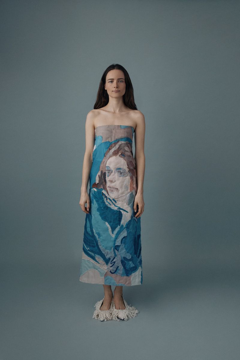 Full-length portrait of a woman with long brown hair wearing a strapless dress with a blue and beige print of a portrait.