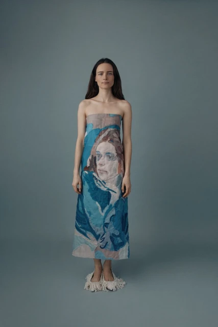 Full-length portrait of a woman with long brown hair wearing a strapless dress with a blue and beige print of a portrait.