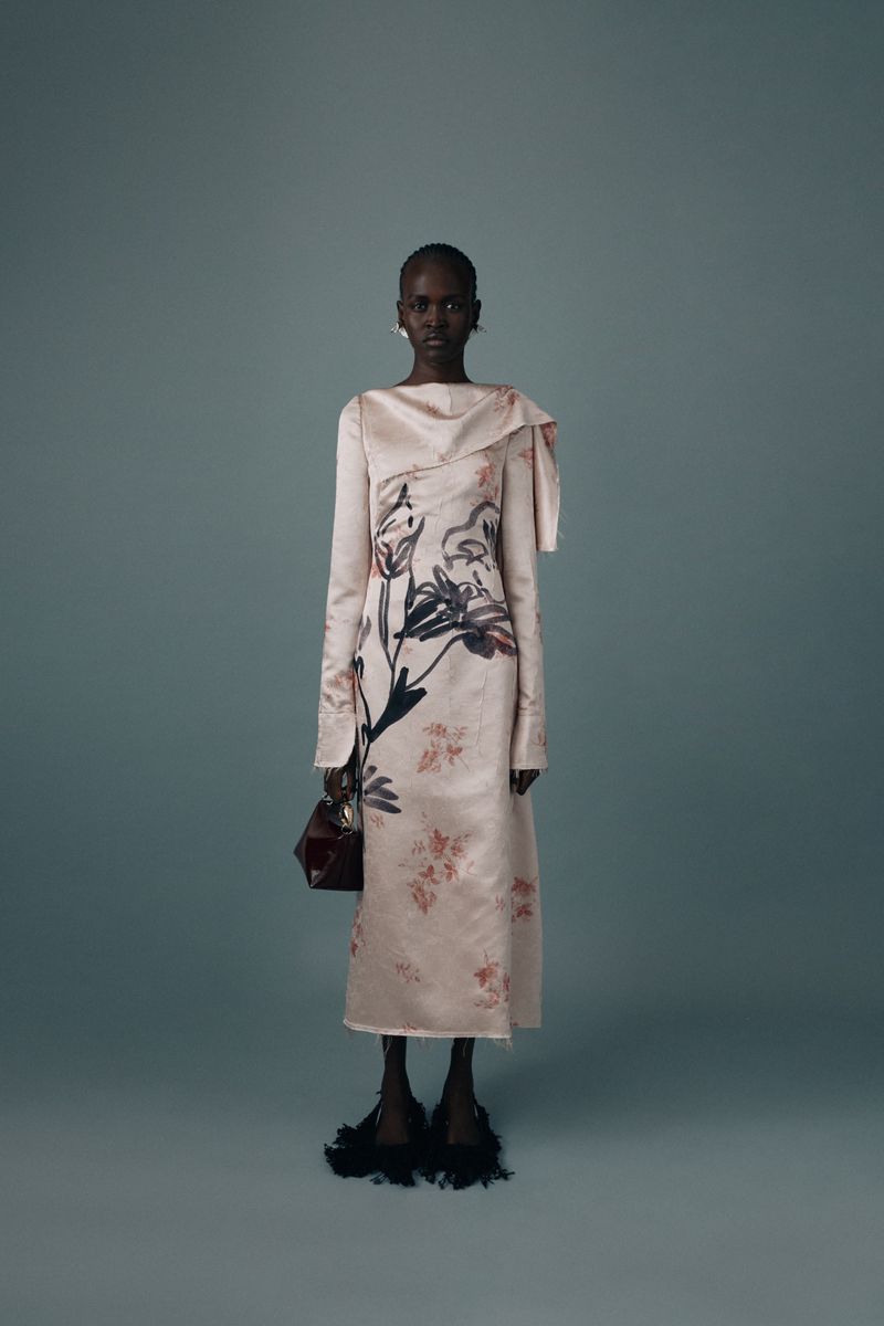 Dark-skinned woman poses in a blush-toned, floor-length gown with a floral print, holding a handbag, against a muted blue-gray background.