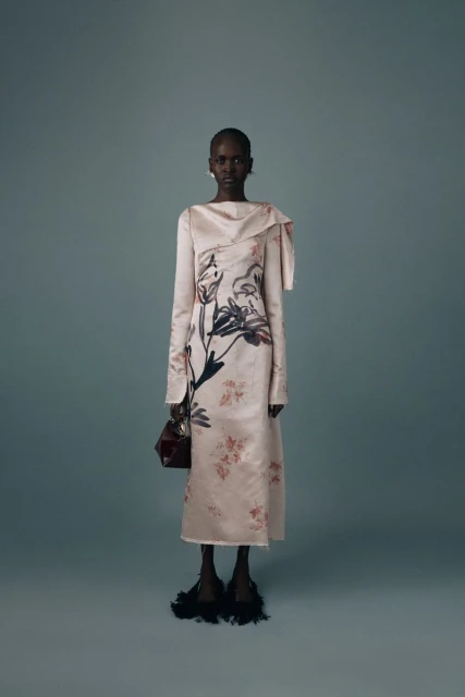 Dark-skinned woman poses in a blush-toned, floor-length gown with a floral print, holding a handbag, against a muted blue-gray background.