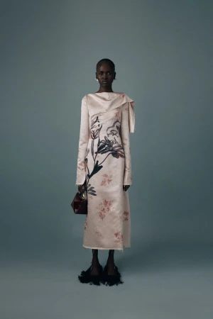 Dark-skinned woman poses in a blush-toned, floor-length gown with a floral print, holding a handbag, against a muted blue-gray background.