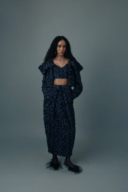 Fashion model posing in textured jacket, bralette, and skirt ensemble with feathered mules against grey backdrop in studio shot.