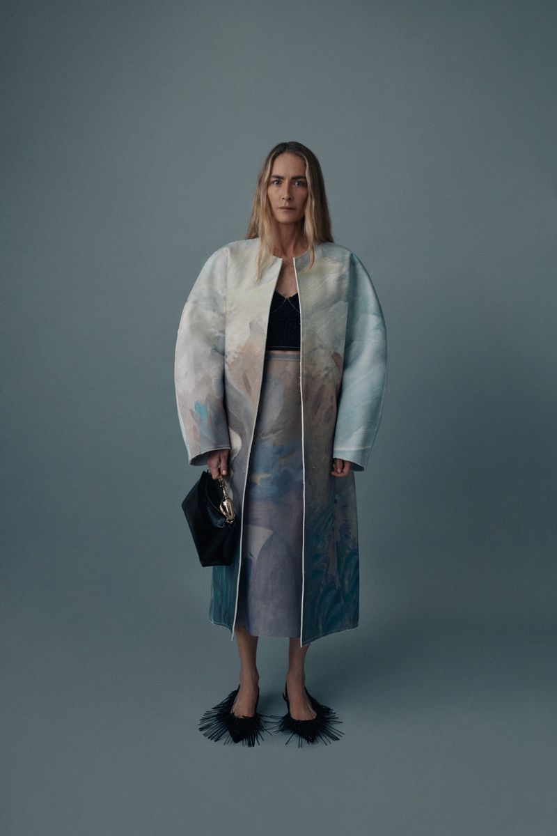 Full-length shot of a blonde adult woman posing in a colorful coat, skirt, black top, and black handbag on a grey studio background.