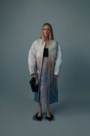Full-length shot of a blonde adult woman posing in a colorful coat, skirt, black top, and black handbag on a grey studio background.