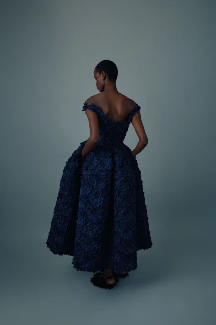 Back view of an African-American woman in an off-the-shoulder, A-line navy gown with textured detailing, standing in a grey studio.