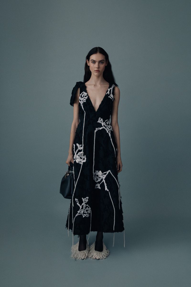 A woman poses in a dark, embroidered dress with a plunging neckline, carrying a black purse, styled against a solid grey background.