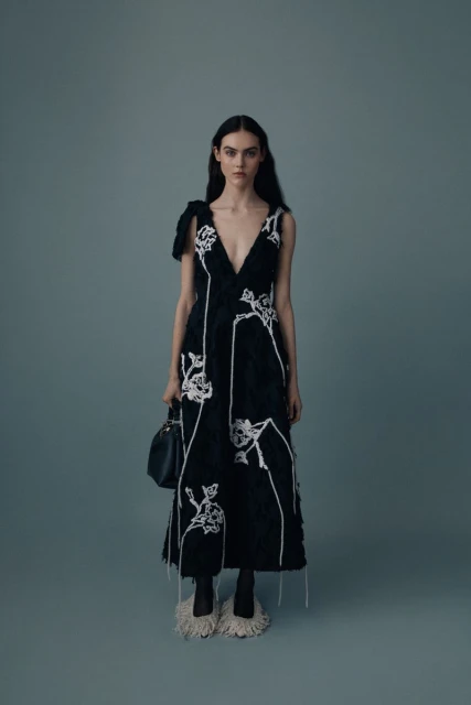 A woman poses in a dark, embroidered dress with a plunging neckline, carrying a black purse, styled against a solid grey background.