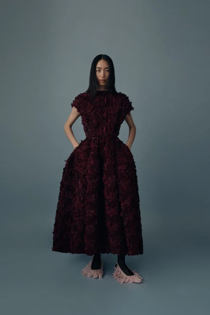 Woman in a textural burgundy dress poses in a studio setting, showcasing fashion with a clean background and soft lighting.