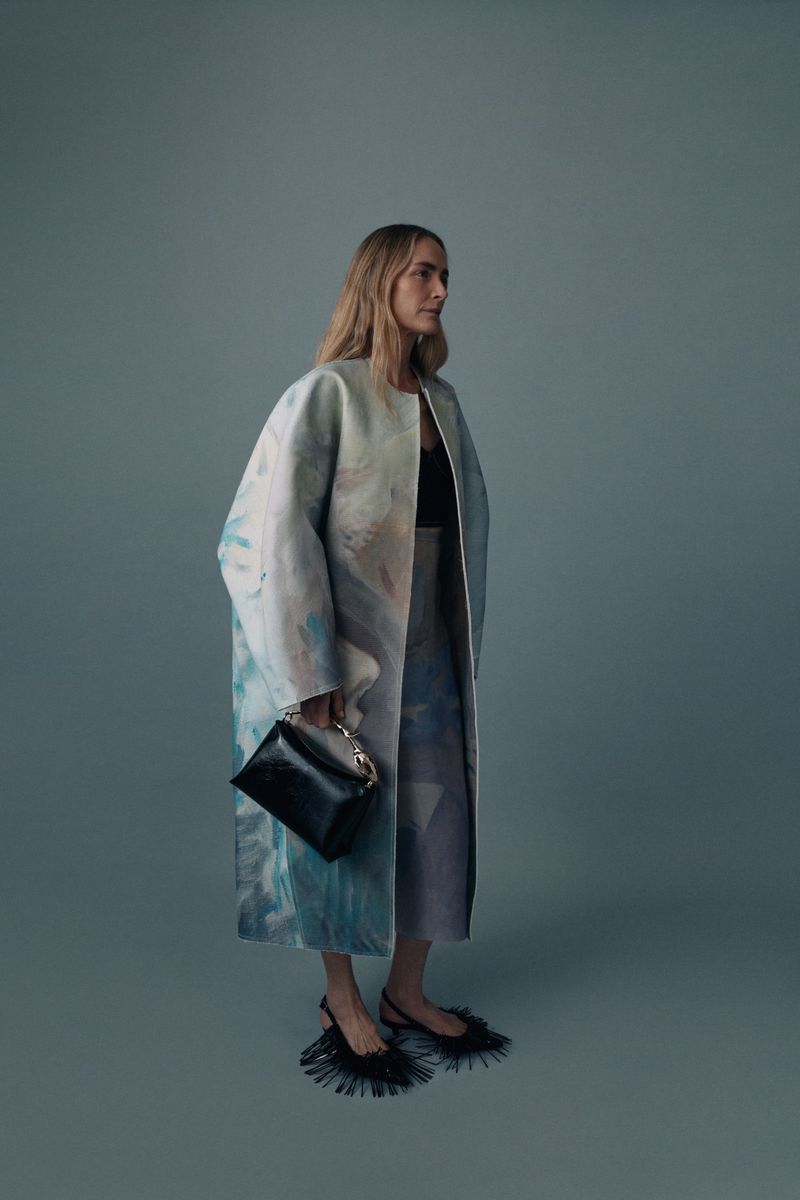 Model wearing an oversized pastel painted coat and black fringe kitten heels while holding a black handbag in a cool blue-grey studio.
