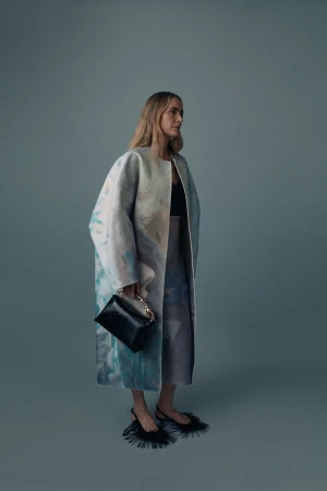 Model wearing an oversized pastel painted coat and black fringe kitten heels while holding a black handbag in a cool blue-grey studio.