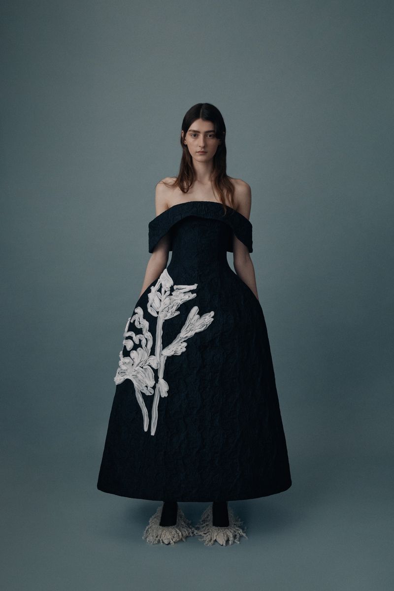 Adult female fashion model wearing a black, off-the-shoulder gown with white floral accents poses in a studio with grey background.