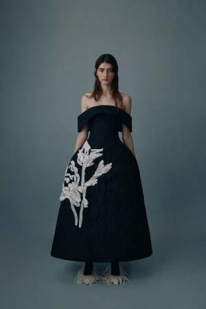 Adult female fashion model wearing a black, off-the-shoulder gown with white floral accents poses in a studio with grey background.