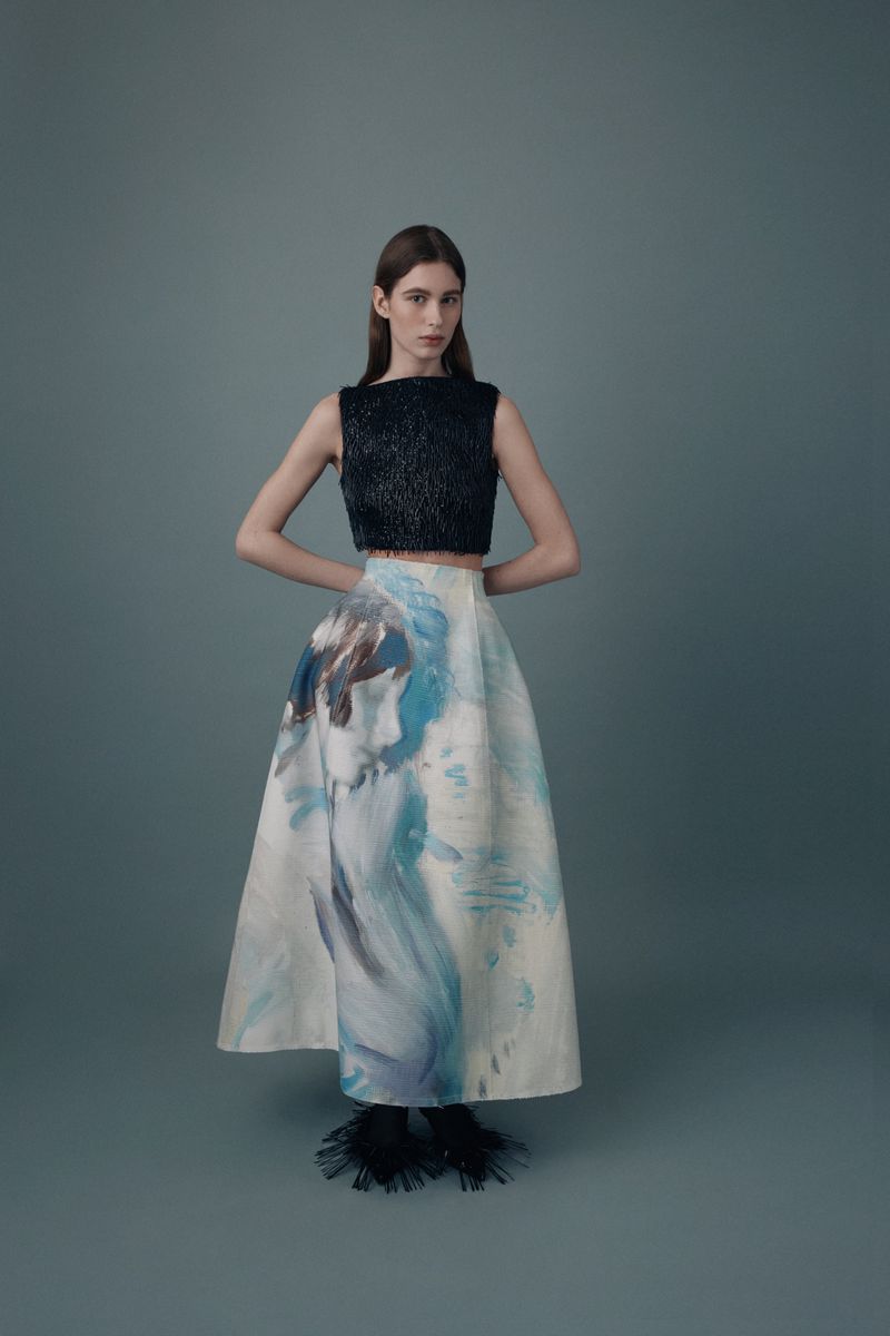 Fashionable woman in a black crop top and artistic white skirt stands in a studio with hands behind her back in a full shot.