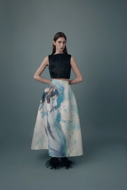 Fashionable woman in a black crop top and artistic white skirt stands in a studio with hands behind her back in a full shot.