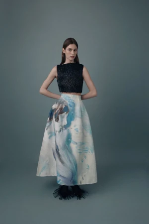 Fashionable woman in a black crop top and artistic white skirt stands in a studio with hands behind her back in a full shot.