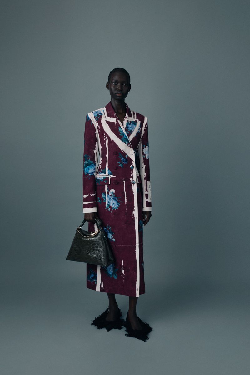 Model wearing a tailored burgundy floral coat with white painted accents and holding a dark handbag against a grey studio backdrop.