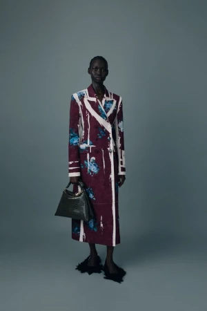 Model wearing a tailored burgundy floral coat with white painted accents and holding a dark handbag against a grey studio backdrop.
