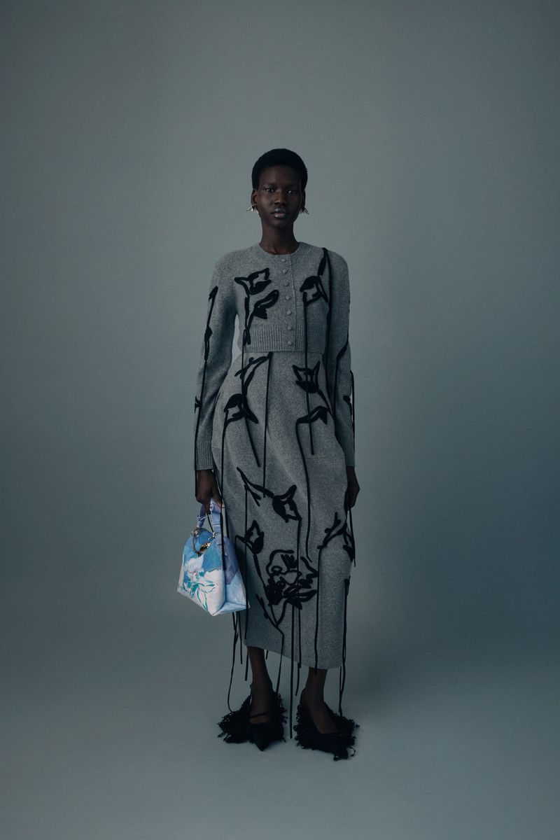 A full-length studio shot of a woman wearing a grey knit dress and cardigan with black floral details, holding a patterned handbag.