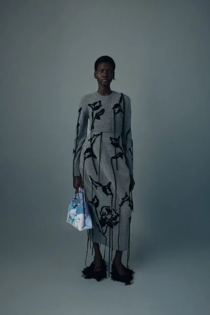 A full-length studio shot of a woman wearing a grey knit dress and cardigan with black floral details, holding a patterned handbag.