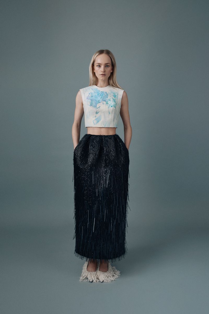 A full shot of a woman in a light colored top with a blue floral design and a dark, fringed, high waisted skirt with light colored shoes.
