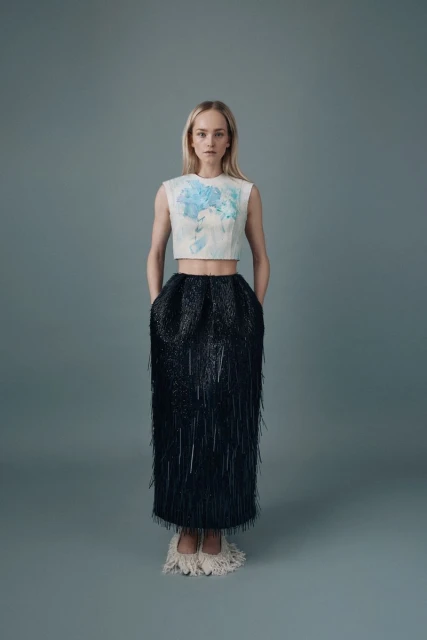 A full shot of a woman in a light colored top with a blue floral design and a dark, fringed, high waisted skirt with light colored shoes.