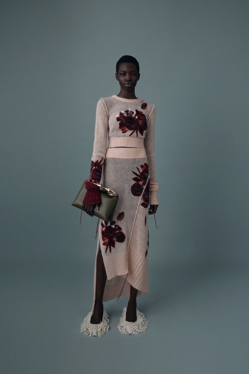 A woman in a sweater and skirt with floral designs stands against a blue background, holding a green clutch.