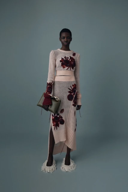 A woman in a sweater and skirt with floral designs stands against a blue background, holding a green clutch.