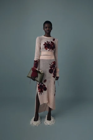 A woman in a sweater and skirt with floral designs stands against a blue background, holding a green clutch.