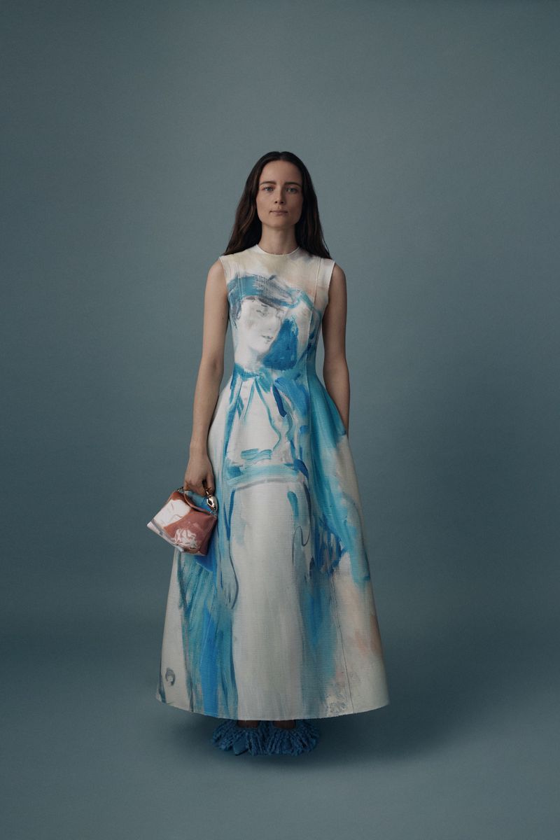 A full shot of a woman in a sleeveless dress with an abstract blue and white pattern, holding a purse against a solid blue background.