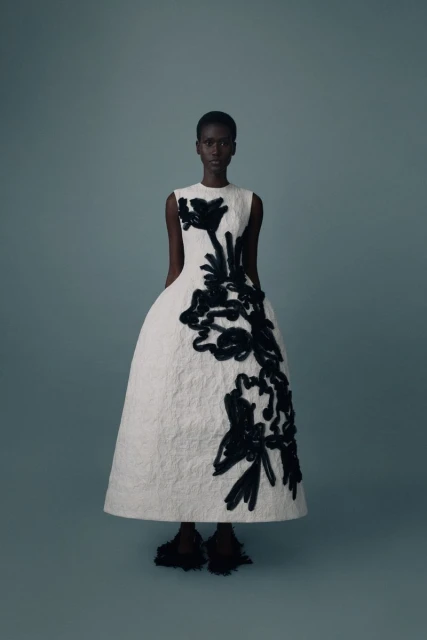 Full-length shot of a Black woman in a formal white A-line dress with large black floral accents on a cool gray background.