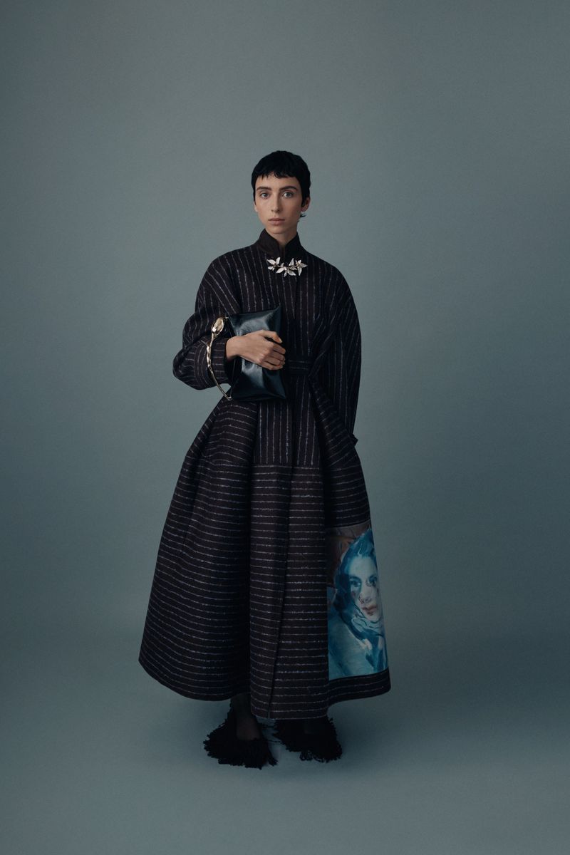 Adult female with short hair poses in a black striped gown holding a clutch purse in a studio with a solid light-blue background.