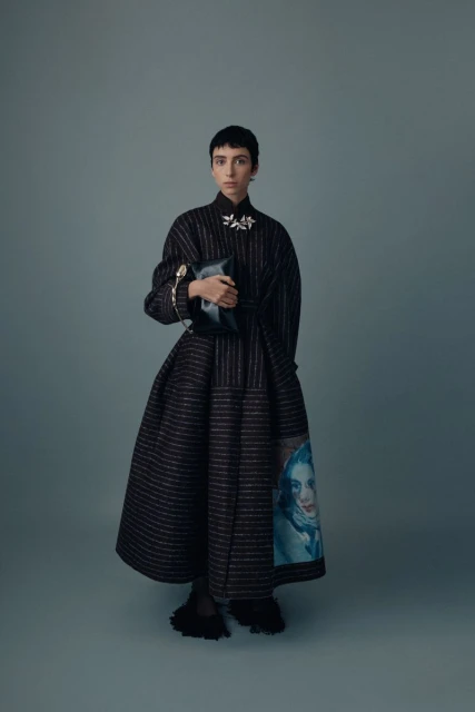 Adult female with short hair poses in a black striped gown holding a clutch purse in a studio with a solid light-blue background.