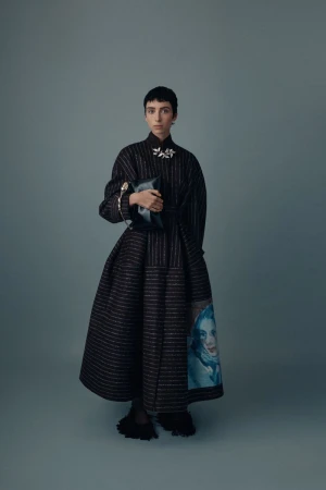 Adult female with short hair poses in a black striped gown holding a clutch purse in a studio with a solid light-blue background.