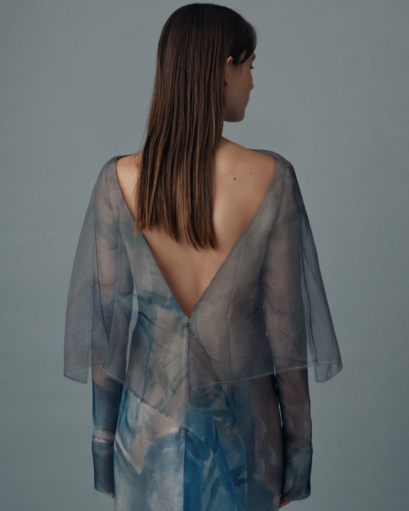 A young woman with long brown hair poses in a studio wearing a semi-sheer blue and gray dress, with her back partially exposed.