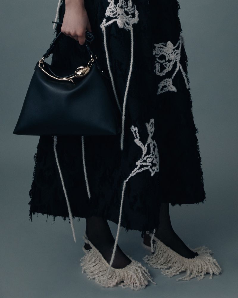 Close-up studio shot of a person wearing a black dress with a white floral pattern, carrying a black handbag, and wearing white fringe shoes.