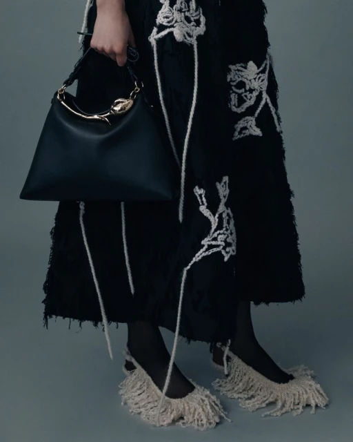 Close-up studio shot of a person wearing a black dress with a white floral pattern, carrying a black handbag, and wearing white fringe shoes.
