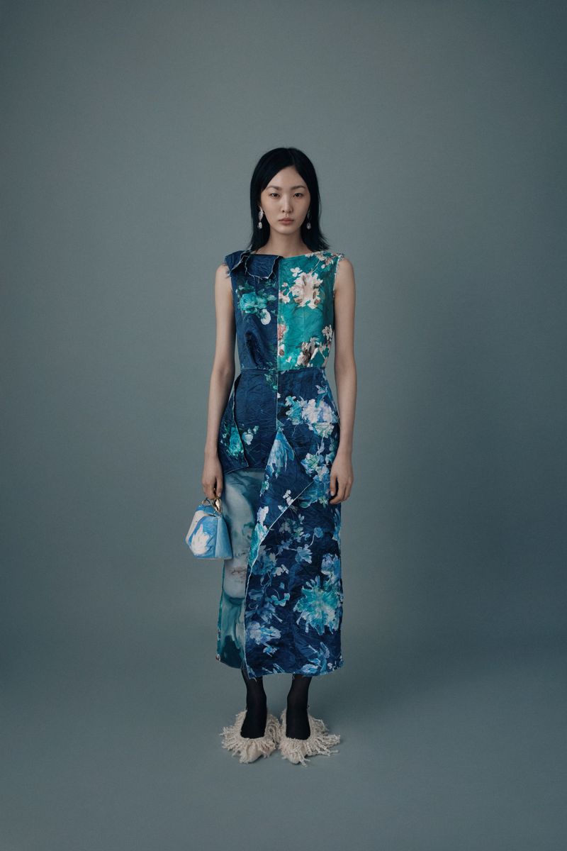 Fashion model poses in a sleeveless floral-print midi dress with a blue patterned handbag on a grey studio backdrop.