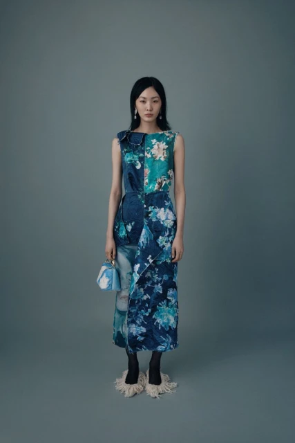 Fashion model poses in a sleeveless floral-print midi dress with a blue patterned handbag on a grey studio backdrop.