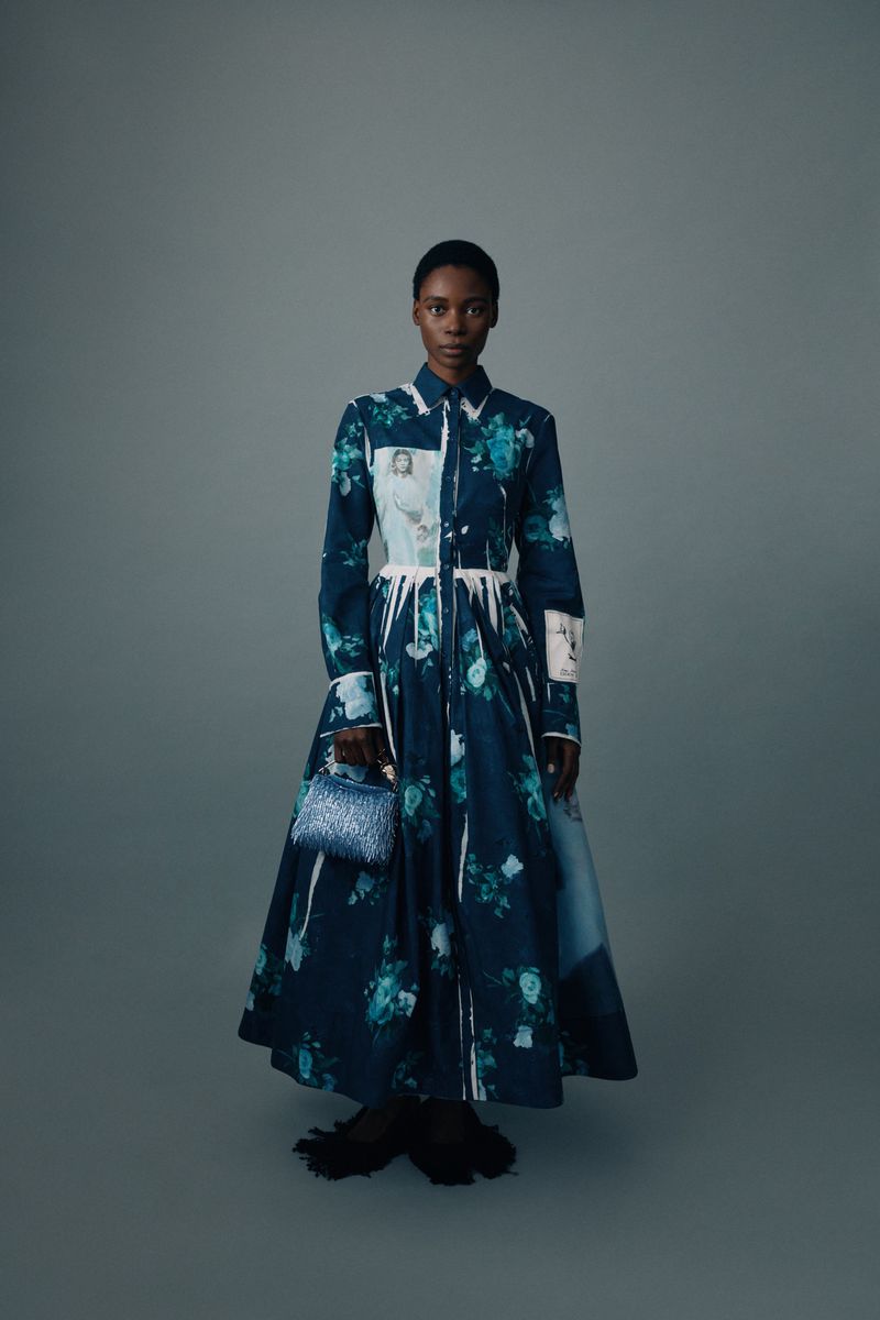 Black female model wearing a printed navy floral collared maxi dress and holding a small handbag against a grey studio backdrop.