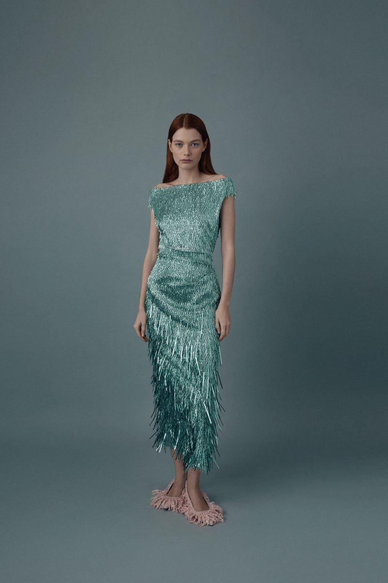 Elegant woman in a shimmering turquoise fringed dress and feathered slippers poses in a studio with a soft, neutral backdrop.