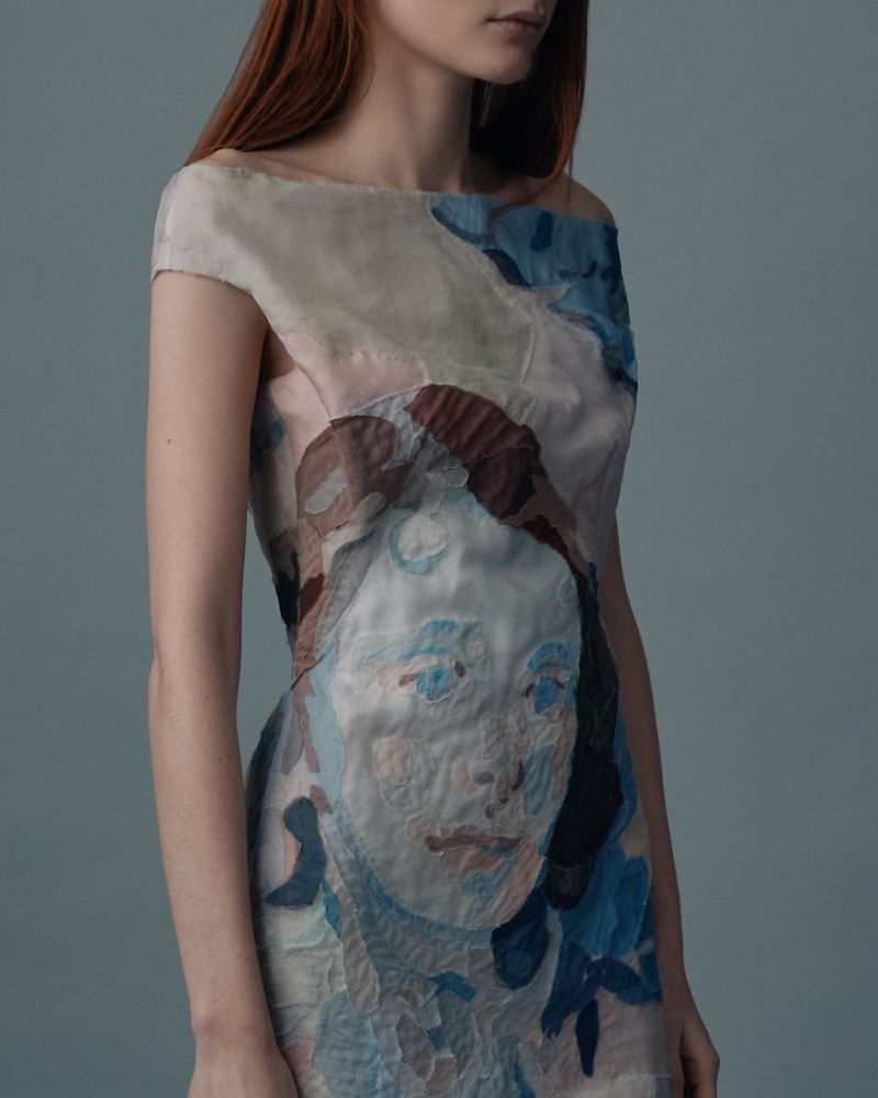 Fashion shot of a red-haired woman wearing a textured dress featuring a portrait, set against a blue-grey backdrop in a studio setting.