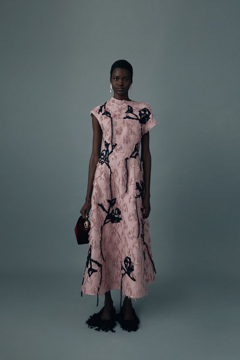 Elegant African-American woman in a pink floral dress poses against a gray backdrop, holding a small red bag and wearing black fringe shoes.