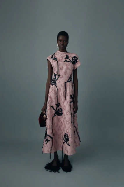 Elegant African-American woman in a pink floral dress poses against a gray backdrop, holding a small red bag and wearing black fringe shoes.