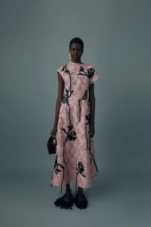 Elegant African-American woman in a pink floral dress poses against a gray backdrop, holding a small red bag and wearing black fringe shoes.