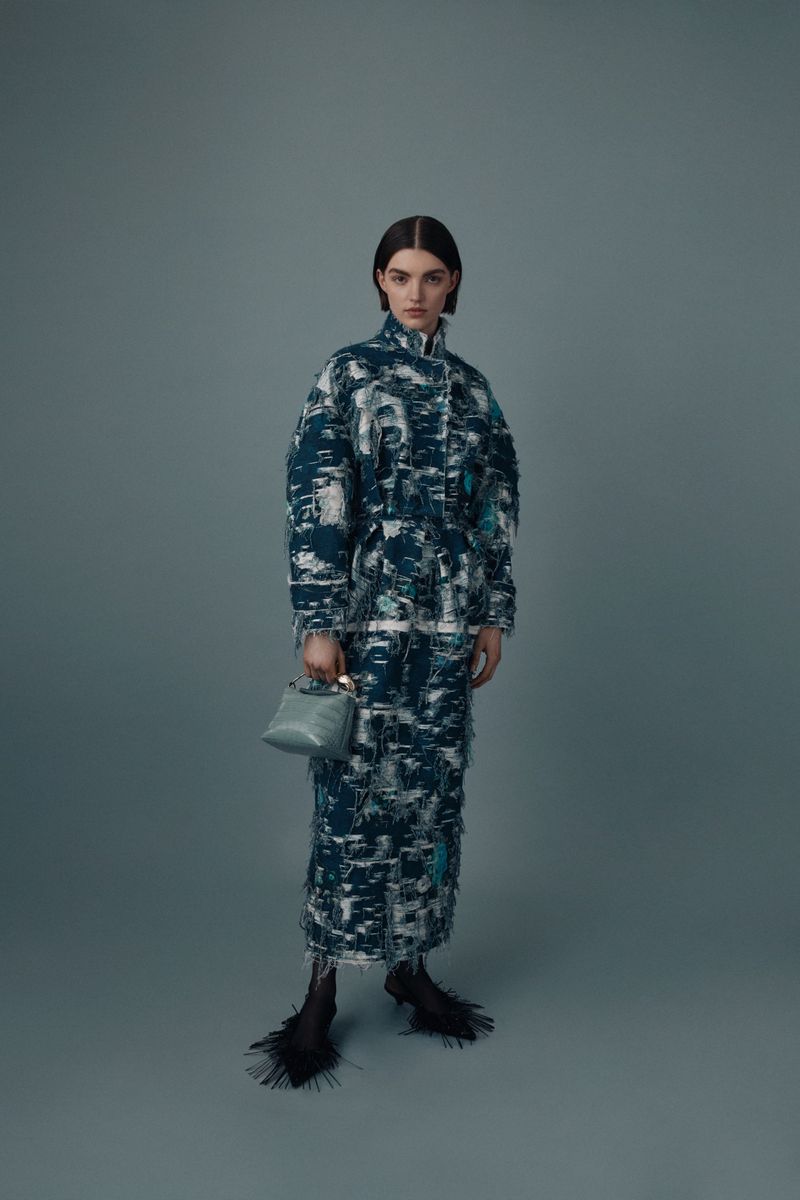 A young woman poses in a full-length shot wearing a long, patterned coat dress and carrying a small blue top-handle handbag in a studio setting.