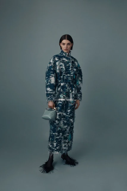 A young woman poses in a full-length shot wearing a long, patterned coat dress and carrying a small blue top-handle handbag in a studio setting.