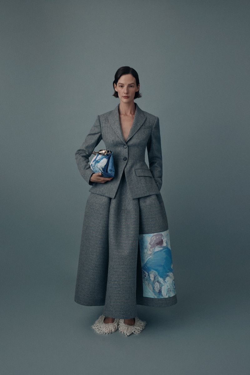 A woman in a grey suit and skirt poses holding a handbag in a studio with a light grey background for a fashion editorial shot.