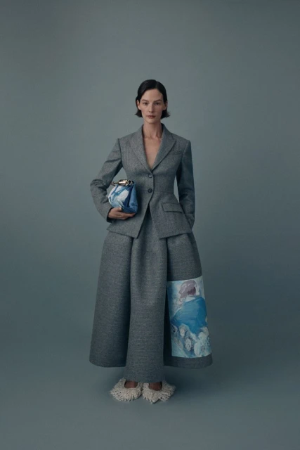 A woman in a grey suit and skirt poses holding a handbag in a studio with a light grey background for a fashion editorial shot.